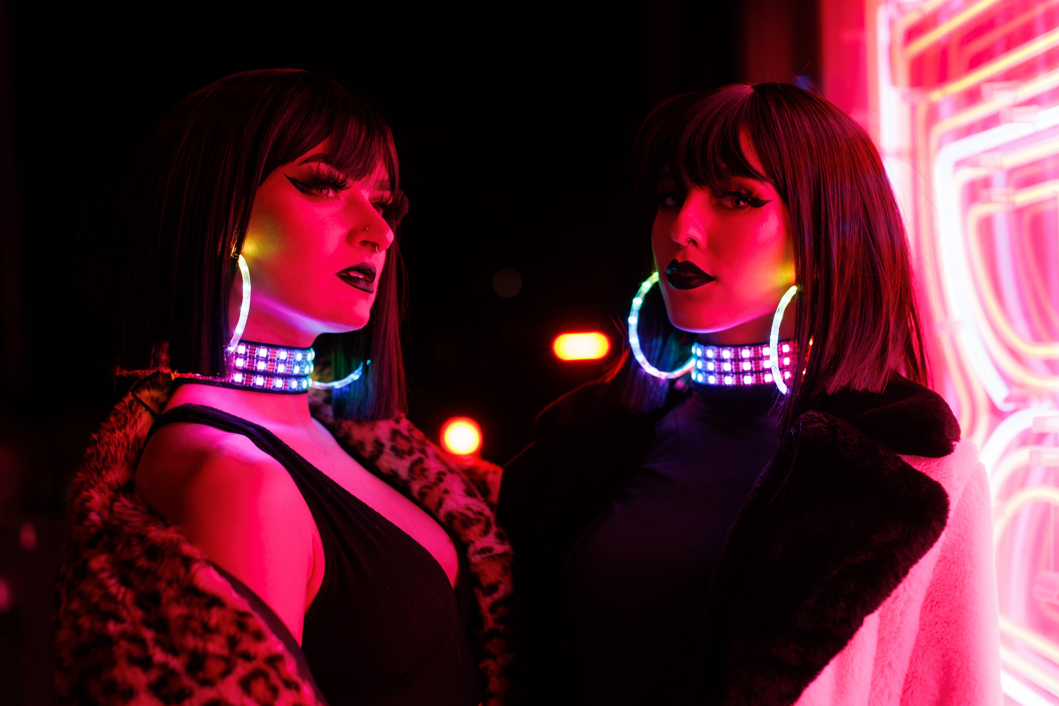 two models wearing led accessories & chic fur coats standing near a pink neon sign. They are wearing bright LED earrings and chokers.